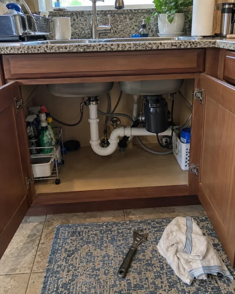 An open kitchen cabinet with under-sink plumbing, a wrench and a towel nearby.
